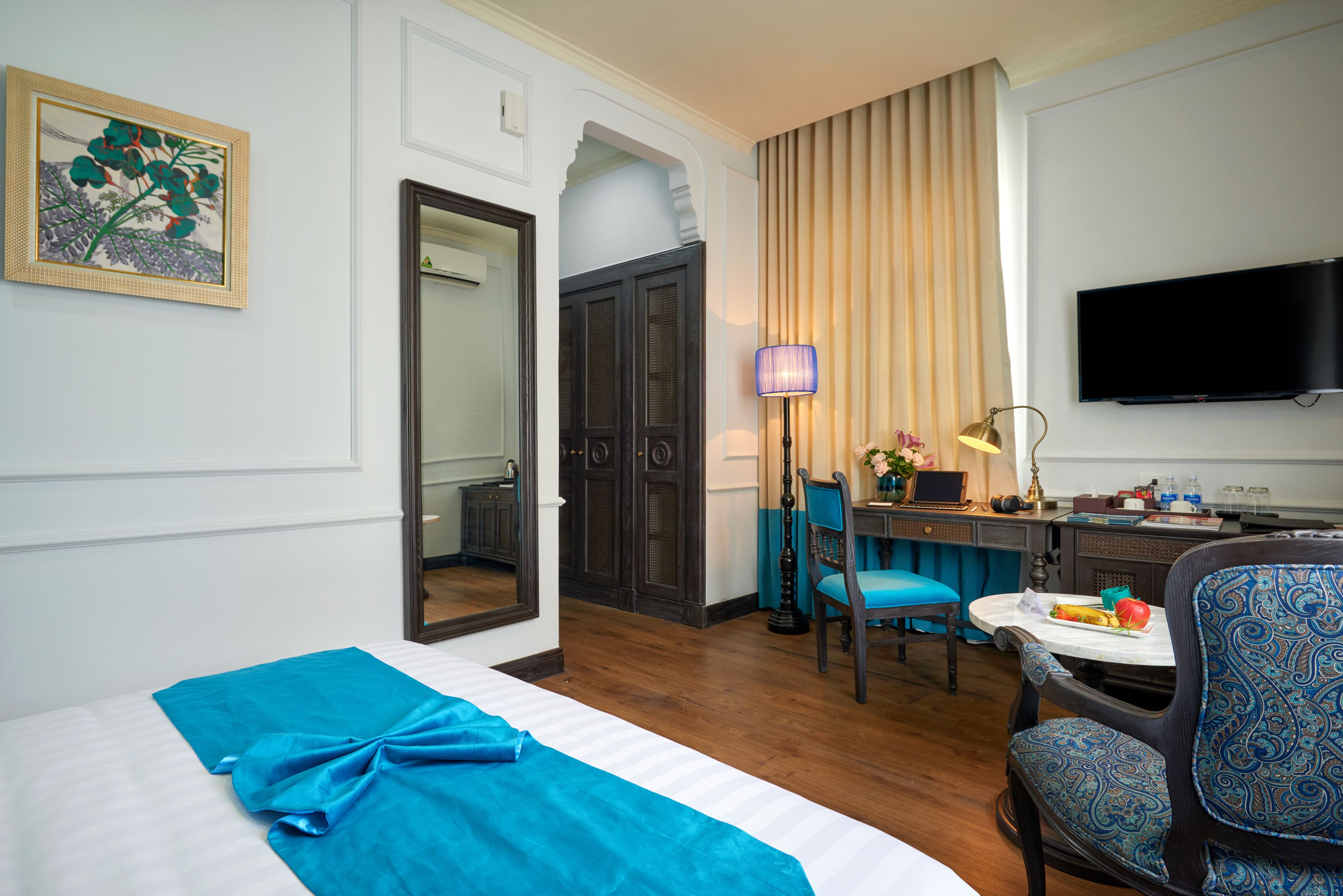 Quarto de hotel com cama, escrivaninha com cadeira, televisão e janela com cortinas.