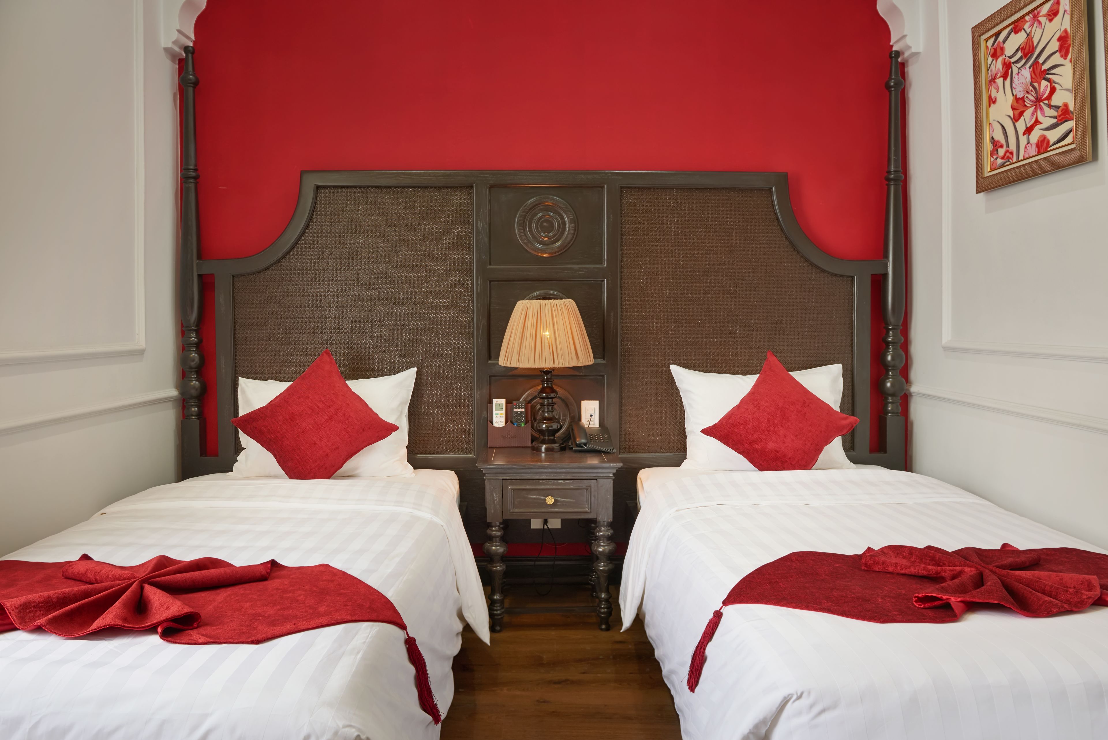 A double room with two beds, each with white bedding and red accents, a wooden headboard, a nightstand with a lamp, and a framed picture on the wall.