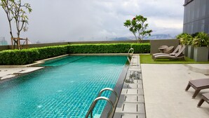 Une piscine avec un bord carrelé, un escalier en métal et des chaises longues sur une terrasse offrant une vue sur la ville.