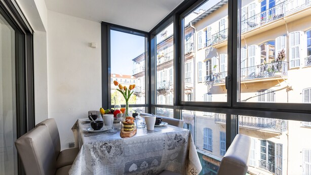 Zona de menjador amb taula preparada per a dues persones, un ram de flors i vista a un edifici residencial a través de grans finestres.