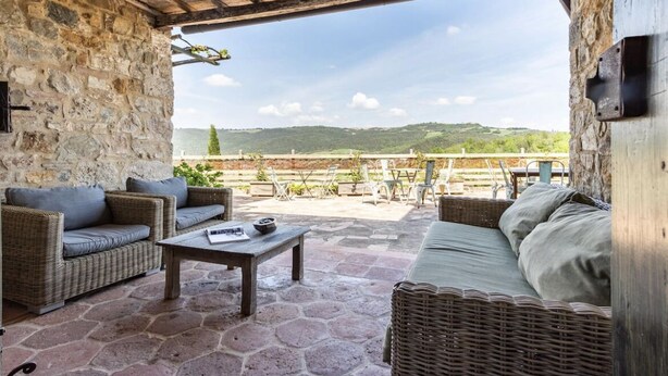 Une terrasse aux murs de pierre, agrémentée de meubles en osier, d’une table et offrant une vue sur un paysage verdoyant.
