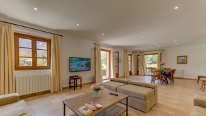 Een ruime woonkamer met een groot raam, een televisie, een eethoek met tafel en stoelen en een bank.