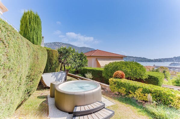 A hot tub with a wooden deck, surrounded by well-manicured hedges and a view of a coastal town. - LA MALMAISON AP1064 by RIVIERA HOLIDAY HOMES (Villefranche-sur-Mer)