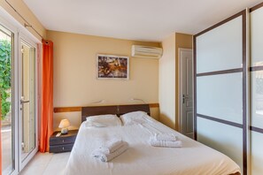 A hotel room with a bed, a nightstand, a lamp, a painting, an air conditioning unit, and a sliding glass door.