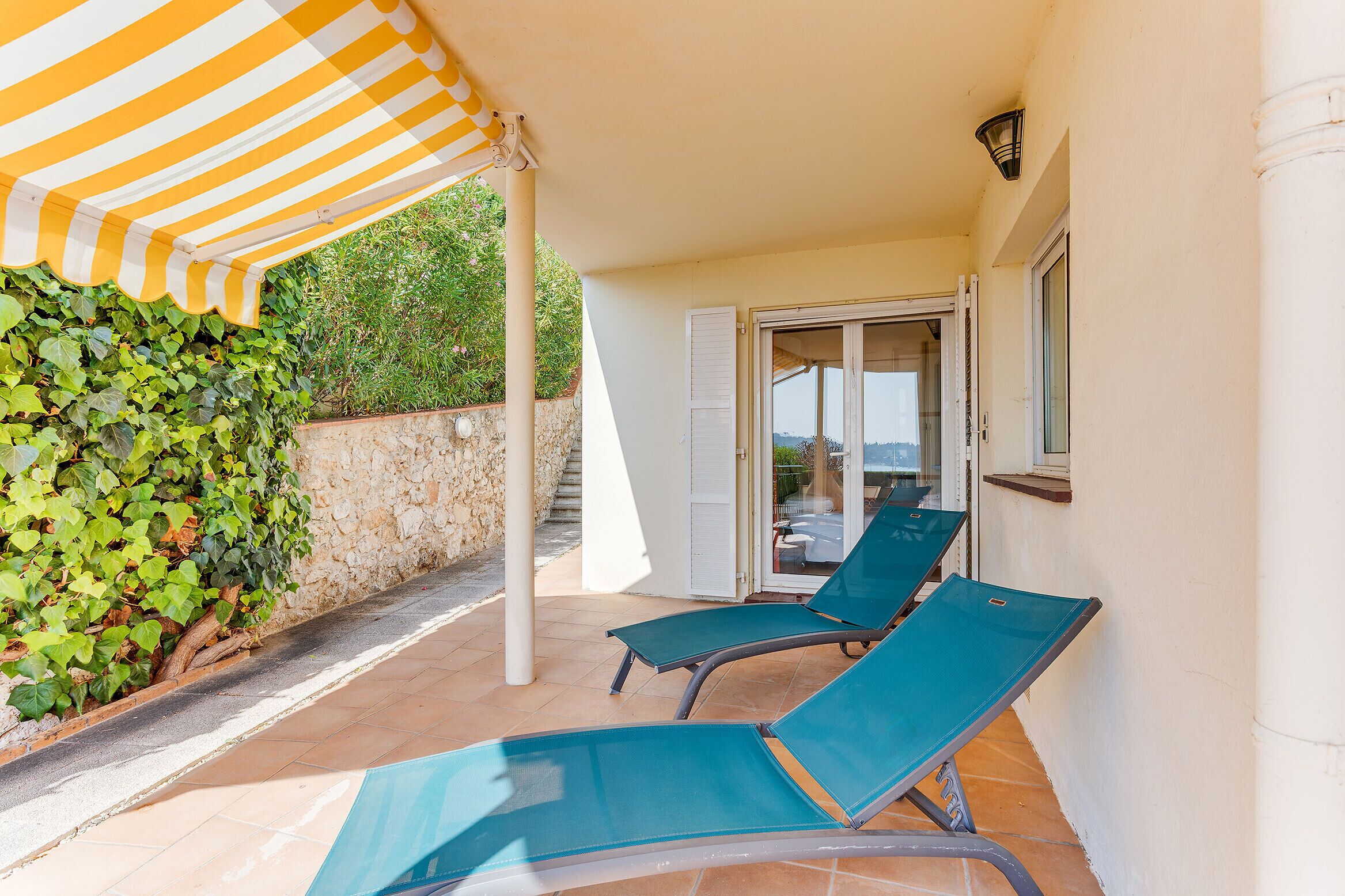 A covered outdoor area with two lounge chairs, a stone wall, and a view of the sea through a glass door.