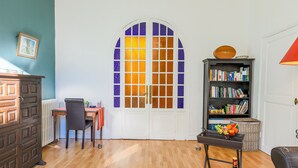 Una habitación con suelo de madera, una mesa de comedor con sillas, una estantería y un amplio ventanal de arco con vidrios de colores.