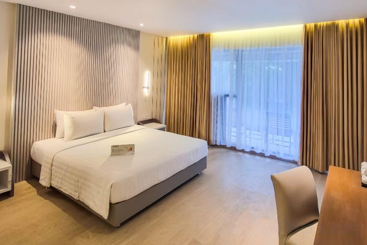 A hotel room with a large bed, bedside tables, a desk, and a view of the outdoors through large windows.