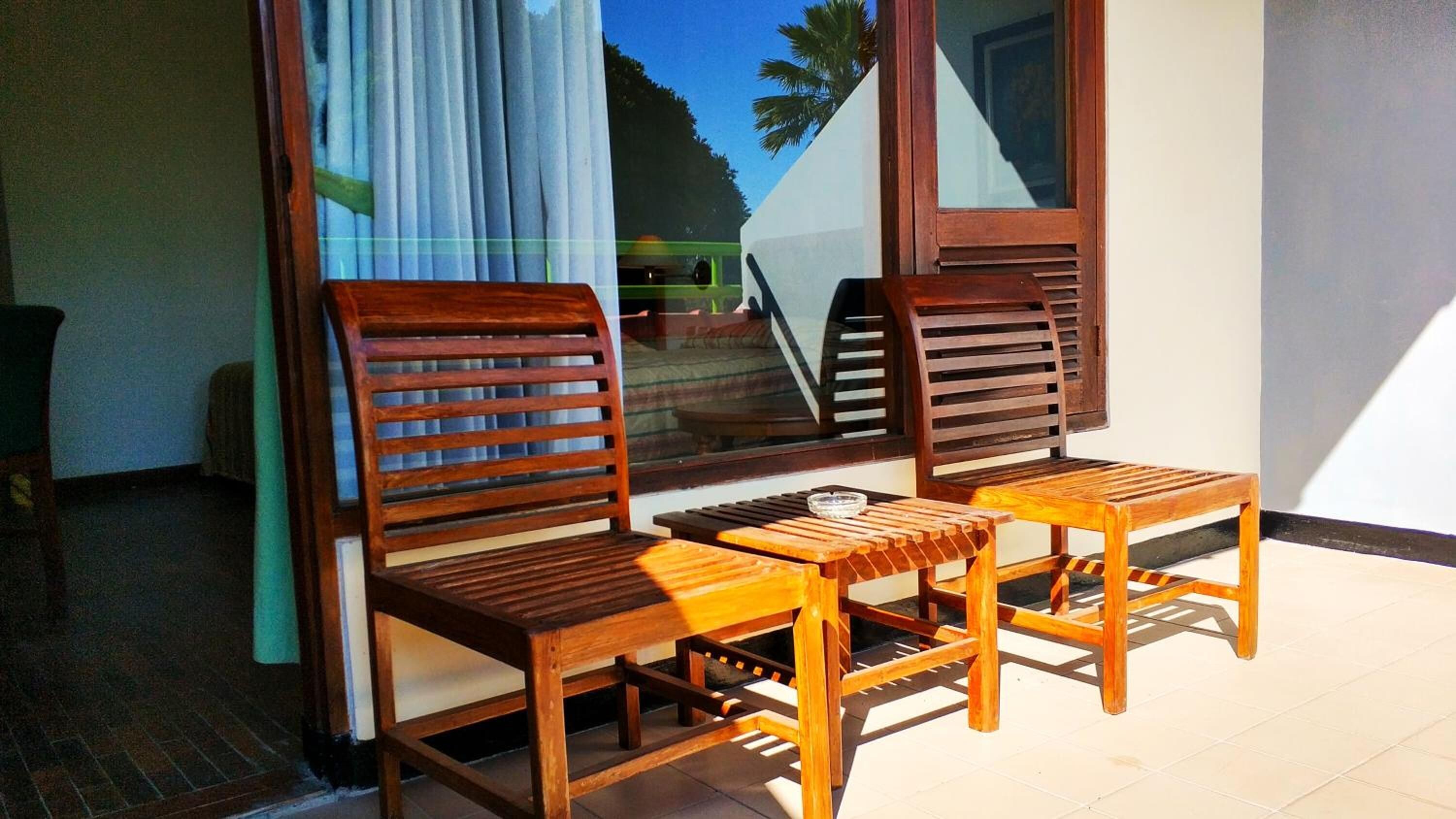 Un espace extérieur aménagé avec des meubles en bois, comprenant deux chaises et une petite table, placé devant une fenêtre donnant sur un escalier et des palmiers.