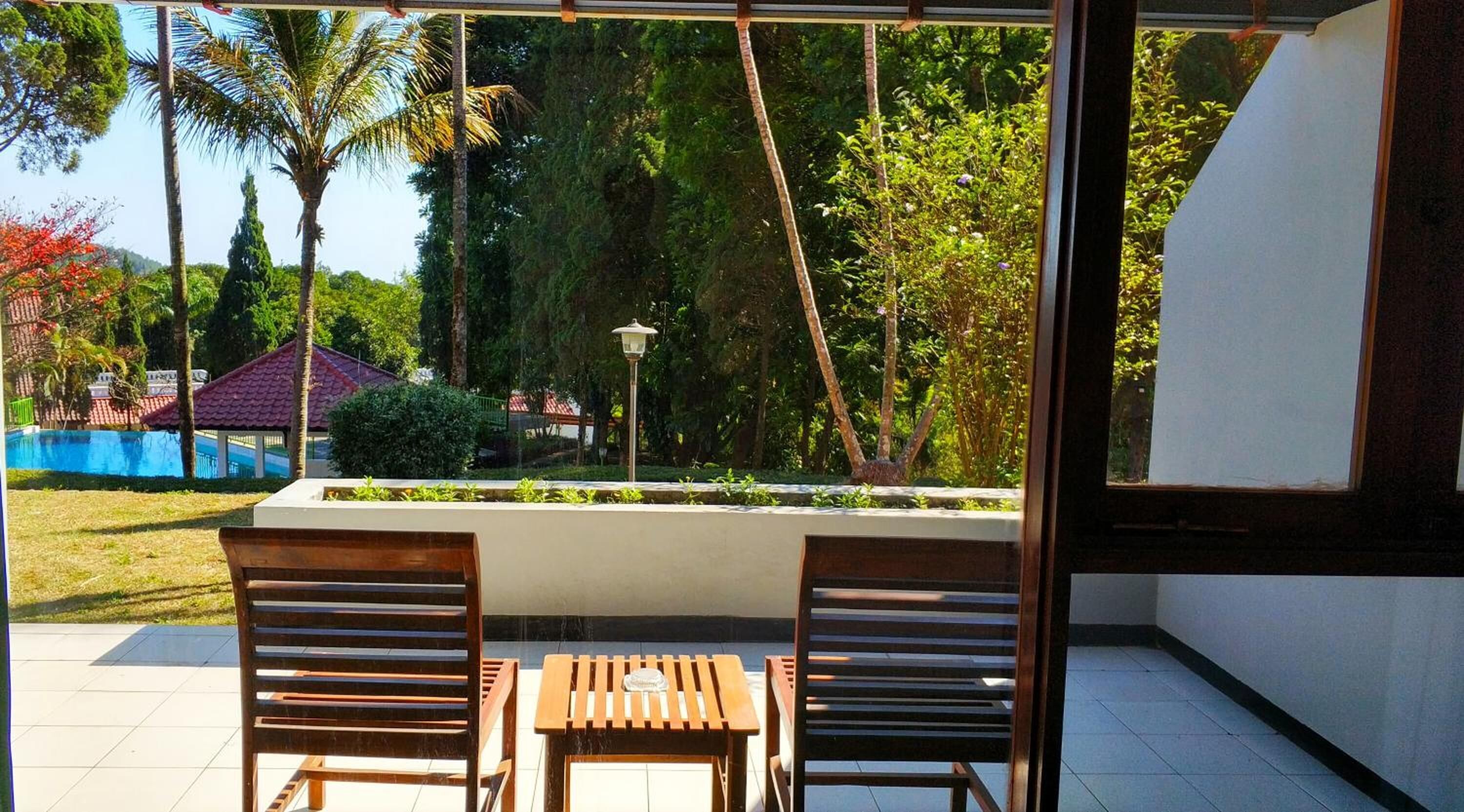 Un patio con dos sillas de madera y una pequeña mesa, con vista a una piscina y abundante vegetación.