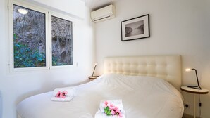 A bedroom with a large bed, a bedside table with a lamp, a framed picture on the wall, and an air conditioning unit. - VILLA HENRIETTA AP4070 by RIVIERA HOLIDAY HOMES (Villefranche-sur-Mer)