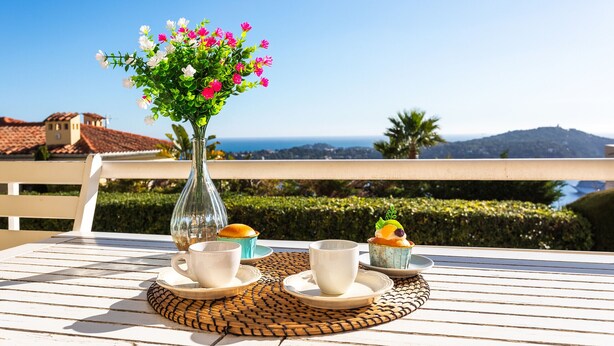 A table with a vase of flowers, two cups, and two pastries on a terrace with a view of the sea and hills. - VILLA HENRIETTA AP4070 by RIVIERA HOLIDAY HOMES (Villefranche-sur-Mer)
