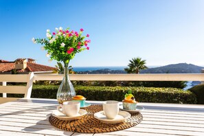 A table with a vase of flowers, two cups, and two pastries on a terrace with a view of the sea and hills. - VILLA HENRIETTA AP4070 by RIVIERA HOLIDAY HOMES (Villefranche-sur-Mer)