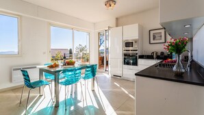 A modern kitchen with white cabinets, a black countertop, and a dining area with blue chairs and a glass table. - VILLA HENRIETTA AP4070 by RIVIERA HOLIDAY HOMES (Villefranche-sur-Mer)