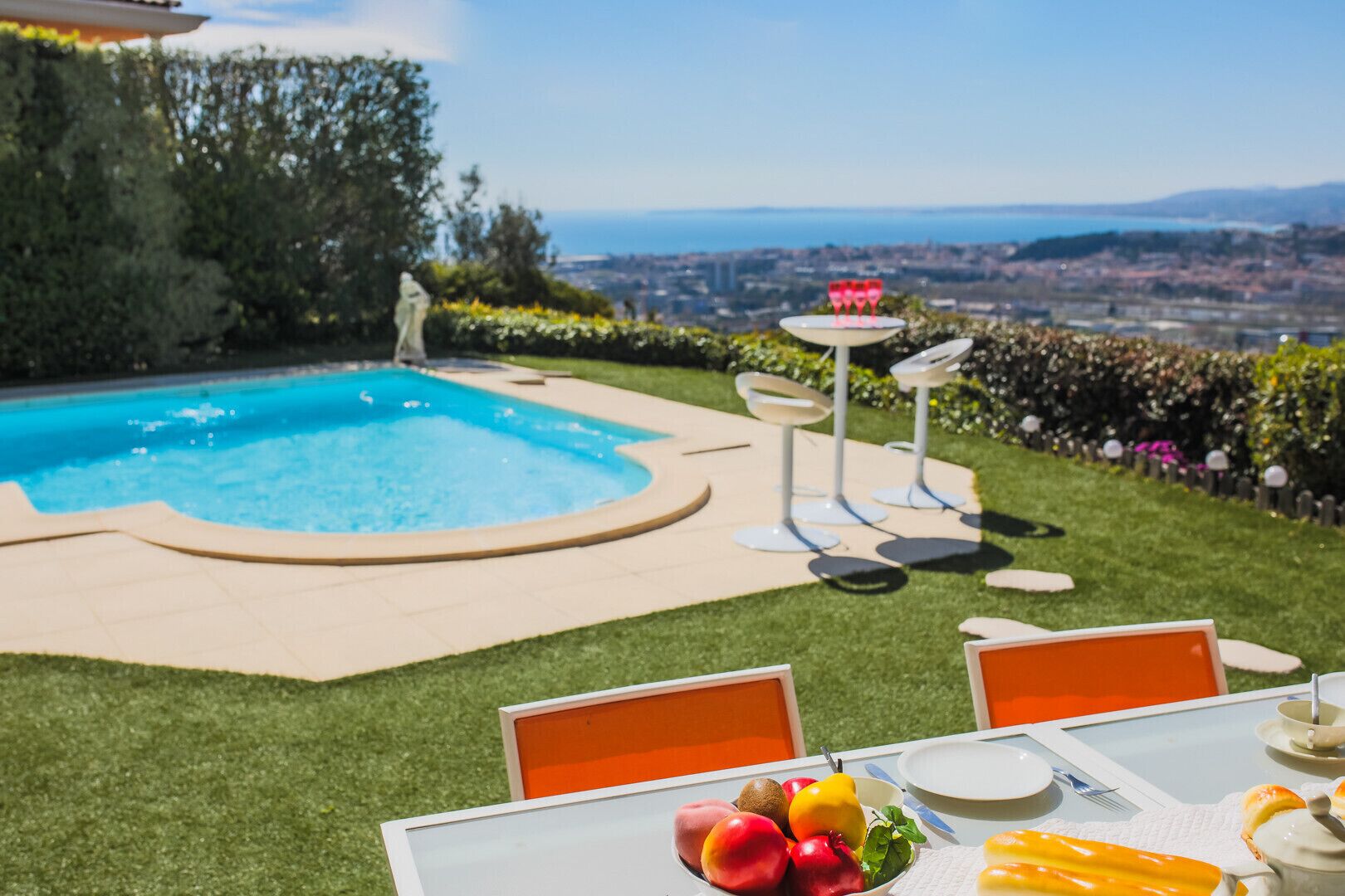 A poolside area with a view of a cityscape, a table with fruit and a glass of water, and modern chairs.