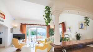 Ein geräumiges Wohnzimmer mit Fernseher, gelb-weißen Sofas, einem Glastisch und Blick ins Freie durch große Fenster.
