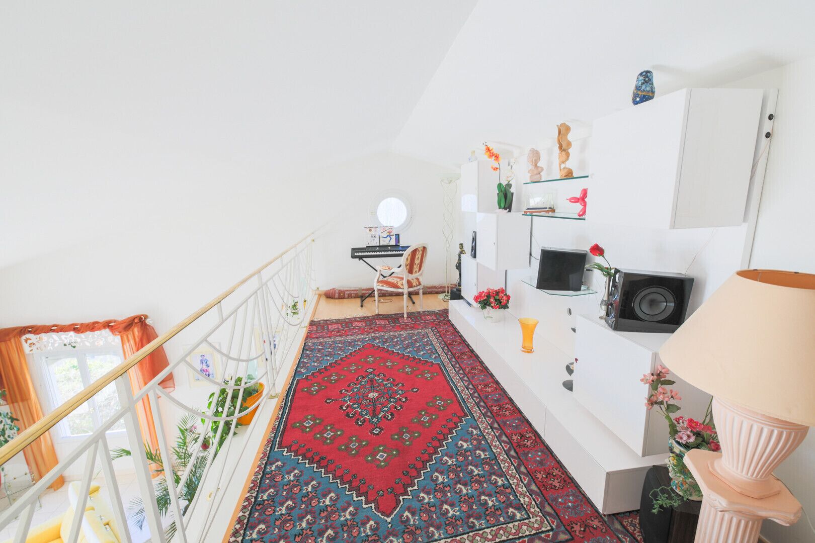 Una habitación amplia con un gran tapete rojo estampado, una consola blanca con objetos decorativos, una silla y un pequeño escritorio con una computadora y una lámpara.
