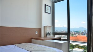 A hotel room with a large window, a bed, a bedside table with a lamp, and a framed picture on the wall.
