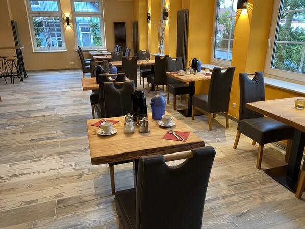 A modern dining area with multiple tables and chairs, a coffee machine, and a view of the outdoors through large windows. - Heidekönig Hotel Celle (Celle)