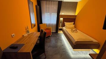 Ein gemütliches Hotelzimmer mit Bett, Schreibtisch, Stuhl, Spiegel, einem Fenster mit Vorhängen und einem kleinen Tisch mit einer Flasche.