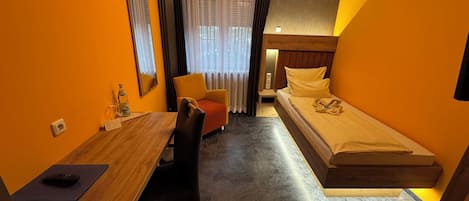 A compact hotel room with a bed, a desk, a chair, a mirror, a window with curtains, and a small table with a bottle.