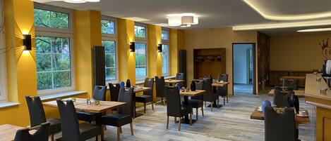 A modern dining area with multiple tables and chairs, large windows with views of greenery, and a bar area with a counter and stools.