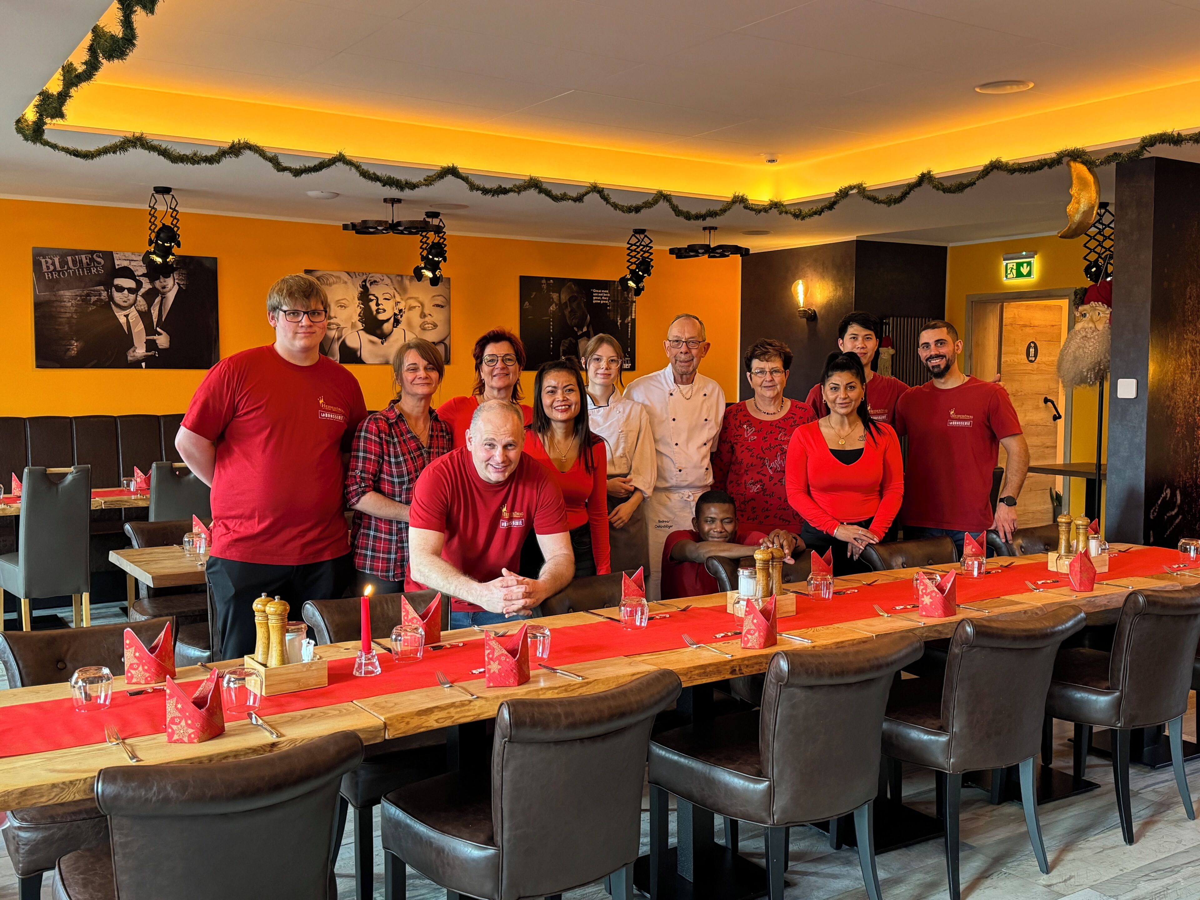 A group of people in a restaurant with a long wooden table, chairs, and a Christmas decoration on the ceiling.