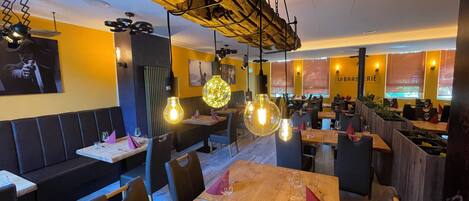 A restaurant interior with wooden tables, black leather booths, hanging pendant lights, and a yellow accent wall.