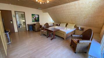 Une pièce spacieuse avec un plancher en bois, un lit recouvert de draps blancs, des chaises en osier, une table à manger et un luminaire suspendu au plafond.