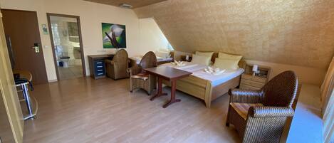 A spacious room with a wooden floor, a bed with white linens, wicker chairs, a dining table, and a ceiling light fixture.