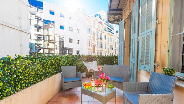 A balcony with wicker furniture, a table with drinks and fruit, and a view of buildings. - CARRE D' OR TERRACE AP4015 by RIVIERA HOLIDAY HOME (Niza)