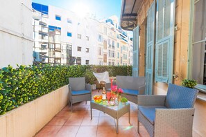 A balcony with wicker furniture, a table with drinks and fruit, and a view of buildings. - CARRE D' OR TERRACE AP4015 by RIVIERA HOLIDAY HOME (Niza)