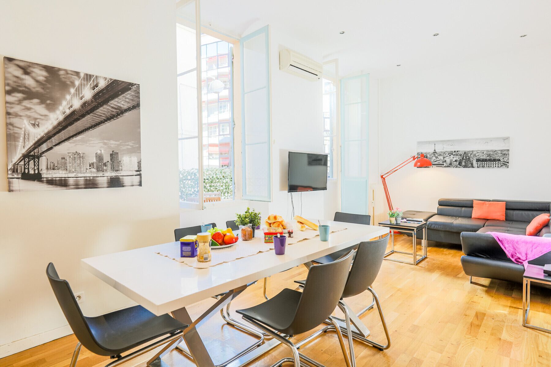 A modern living room with a dining area, featuring a white table, black chairs, a red floor lamp, and a black sofa with orange cushions.