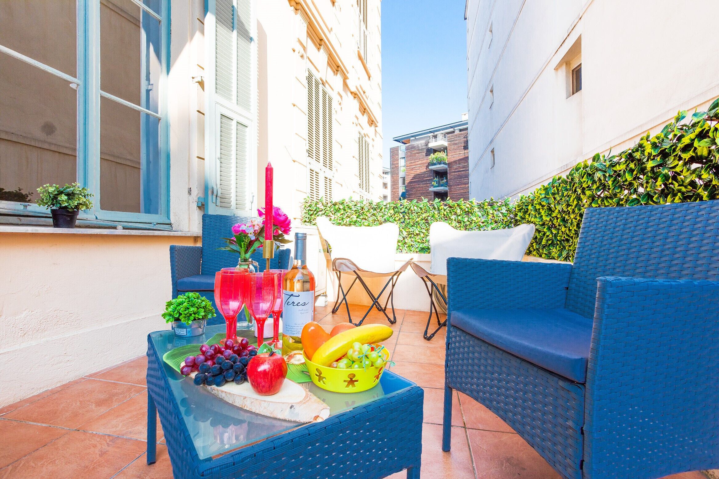 A small outdoor seating area with wicker furniture, a glass table with fruit and drinks, and a potted plant.