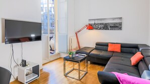 A modern living room with a flat-screen TV, a black leather sofa with orange cushions, a small table with a lamp, and a cityscape painting on the wall. - CARRE D' OR TERRACE AP4015 by RIVIERA HOLIDAY HOME (Niza)