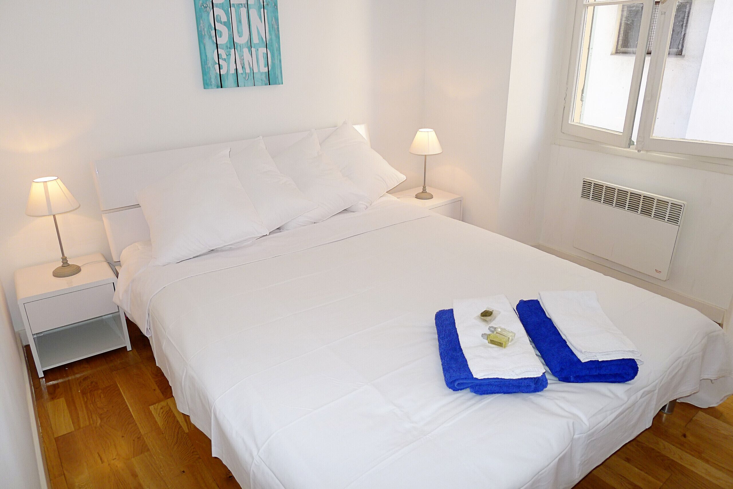 A neatly made bed with white linens, two bedside lamps, a small nightstand, and a window with a view of the outside.