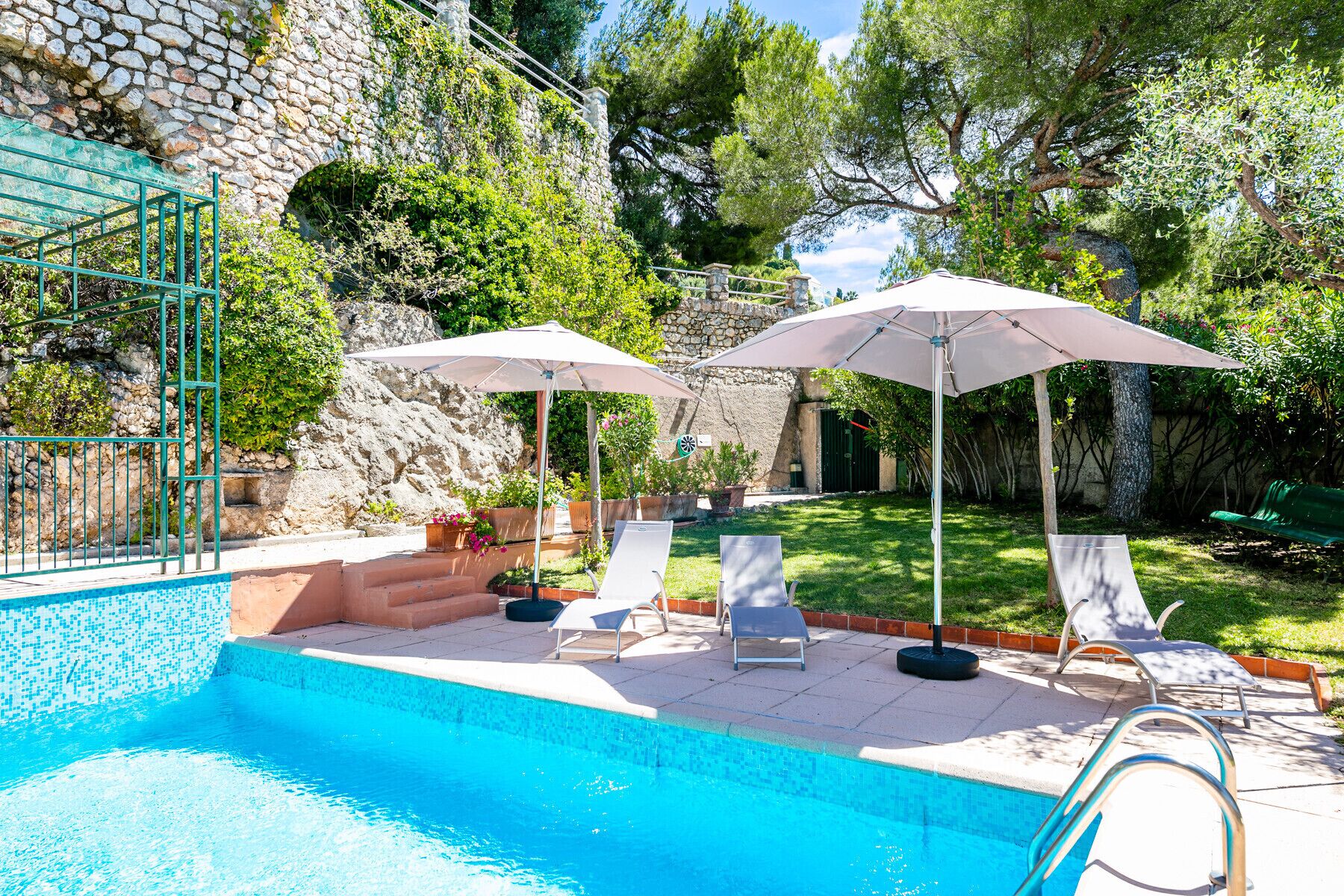 A swimming pool with lounge chairs and umbrellas, surrounded by a stone wall and greenery.