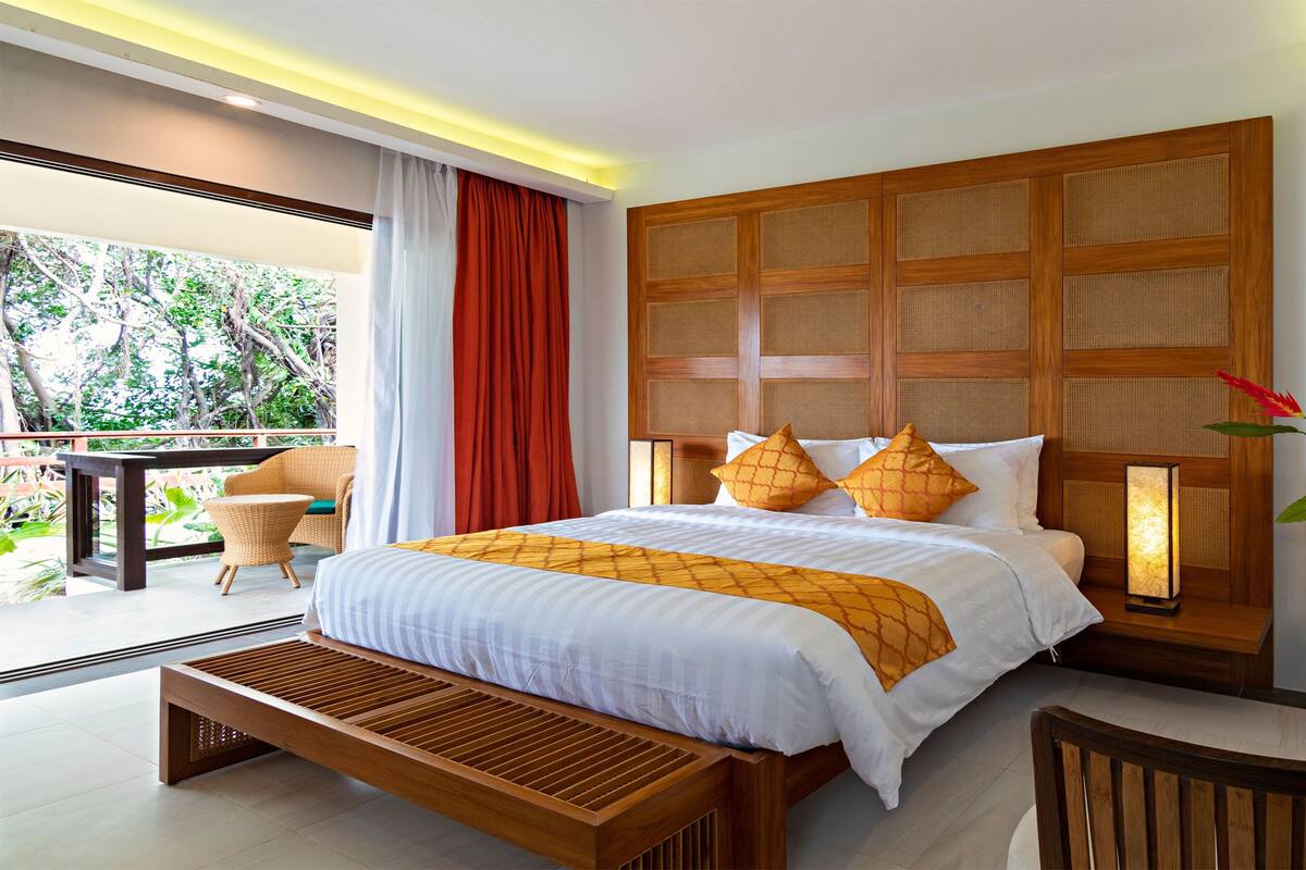 A modern hotel room with a large bed, wooden headboard, and a balcony with wicker furniture.