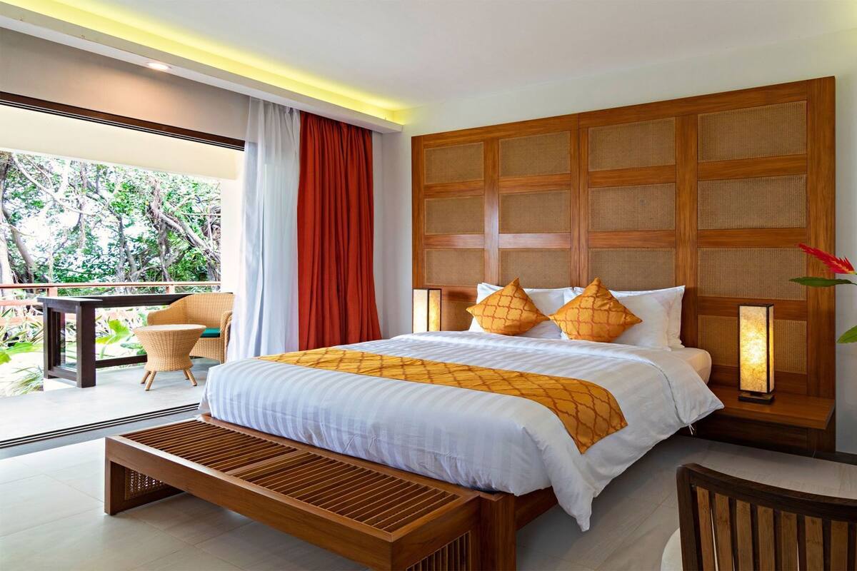 A modern hotel room with a large bed, a wooden headboard, a balcony with a view of greenery, and a wooden bench.