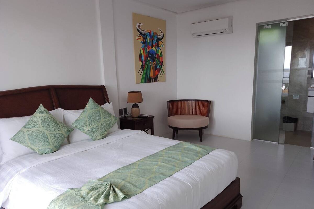 A hotel room with a large bed, a chair, a nightstand, a wall-mounted air conditioner, and a glass door leading to another room.