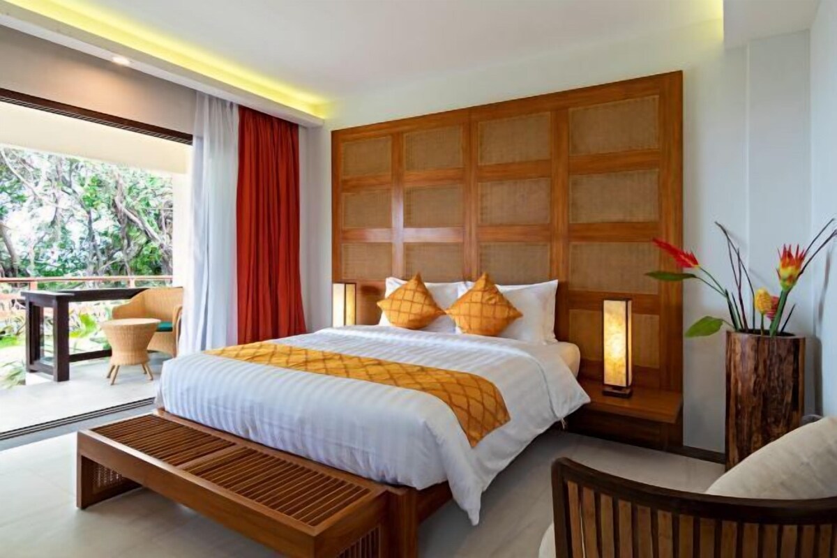 A hotel room with a large bed, a wooden headboard, a bedside table with a lamp, a bench, and a view of the outdoors.