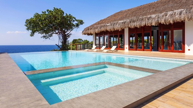 Un espace aménagé au bord de la piscine, avec vue sur l’océan, un bâtiment à toit de chaume et des chaises longues.