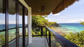Un balcon doté de portes vitrées donnant sur une plage tropicale, avec une eau turquoise et limpide et des formations rocheuses.