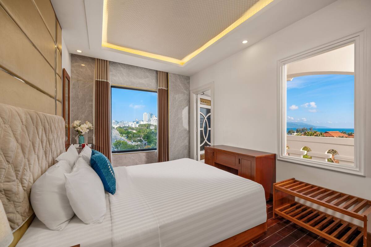 A hotel room with a large bed, a wooden bench, a view of the city, and a window with a balcony.