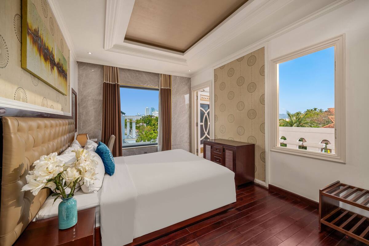 A hotel room with a large bed, a vase of flowers, a view of the city, and a balcony with a railing.