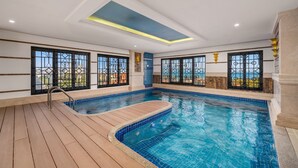 A large indoor swimming pool with a wooden deck, large windows with metal bars, and a skylight.