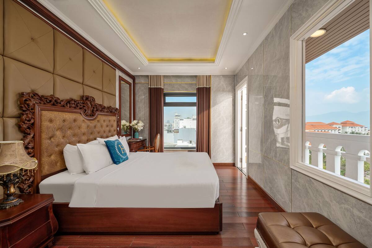 A spacious bedroom with a large bed, a wooden nightstand, and a view of the city through a large window.