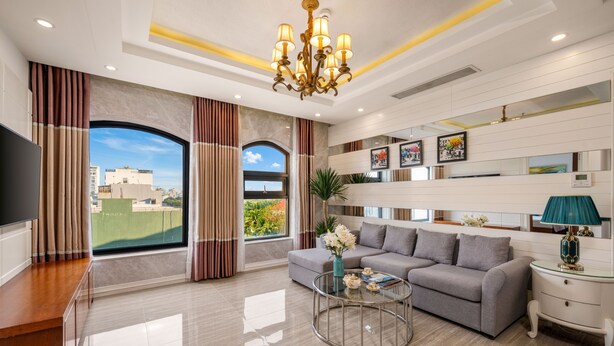 A modern living room with a grey sofa, a coffee table, and a TV on a wooden stand. There is a large window with curtains, a chandelier, and wall-mounted shelves with pictures.