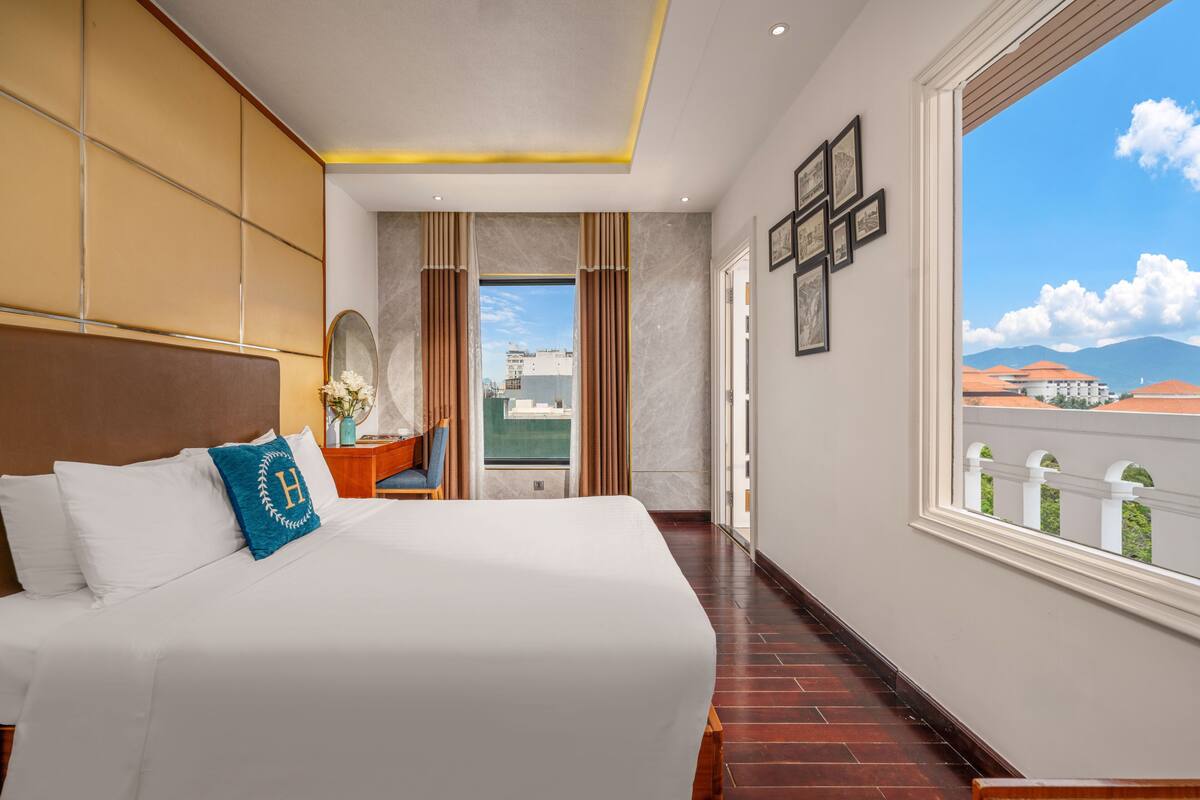 A hotel room with a large bed, a desk with a chair, a large window with a view of buildings and mountains, and a cross on the wall.
