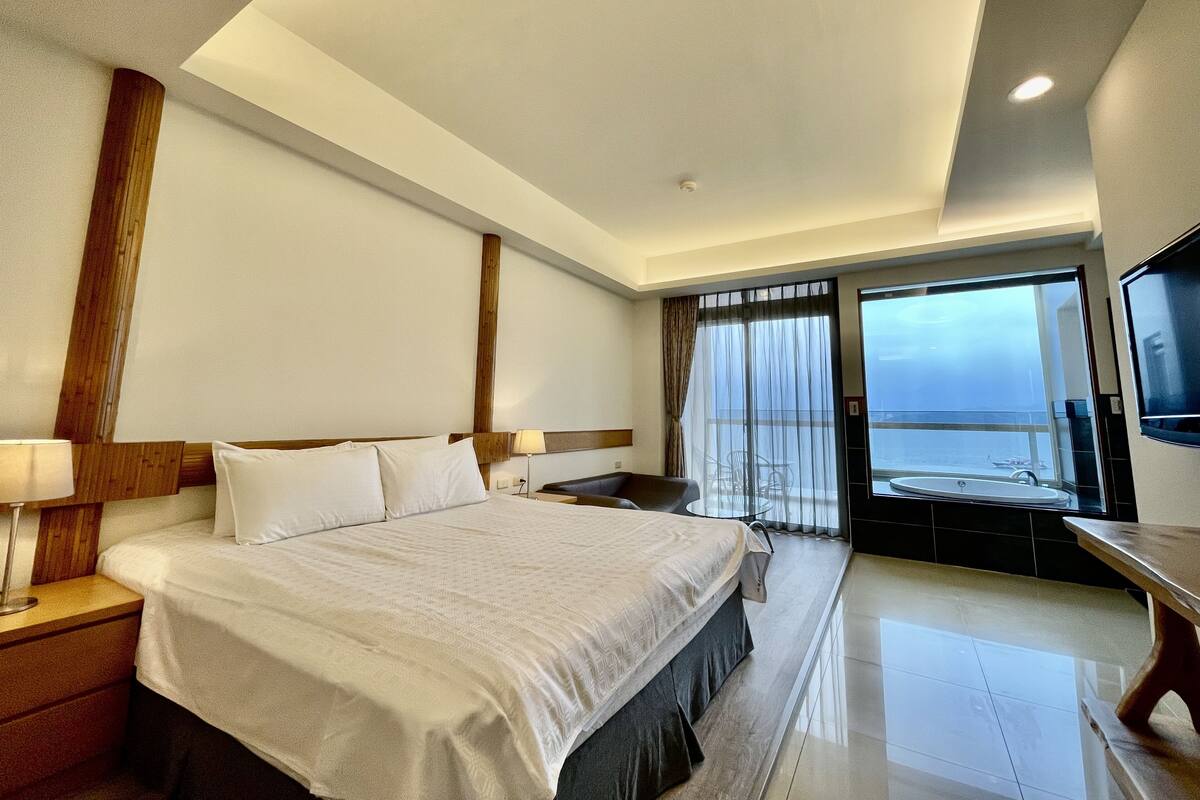 Habitación de hotel con una cama grande, un escritorio, una silla, un televisor y vista al mar a través de un ventanal amplio.