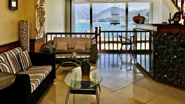 A spacious living area with a sofa, chairs, and a glass table. A large window offers a view of the sea and mountains. - Ming Hu B&B (No elevator) (Yuchi)